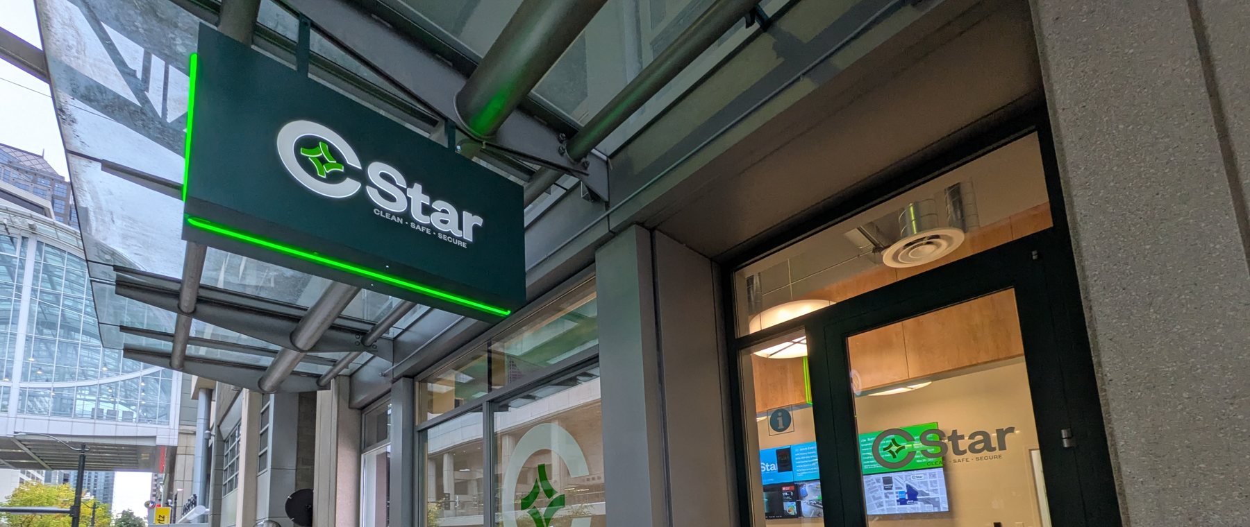 The C-STAR storefront at 810 Pike, its green "CStar · Clean · Safe · Secure" blade sign lit under the glass canopy, its glass door looking in on a reception counter and wall screens.